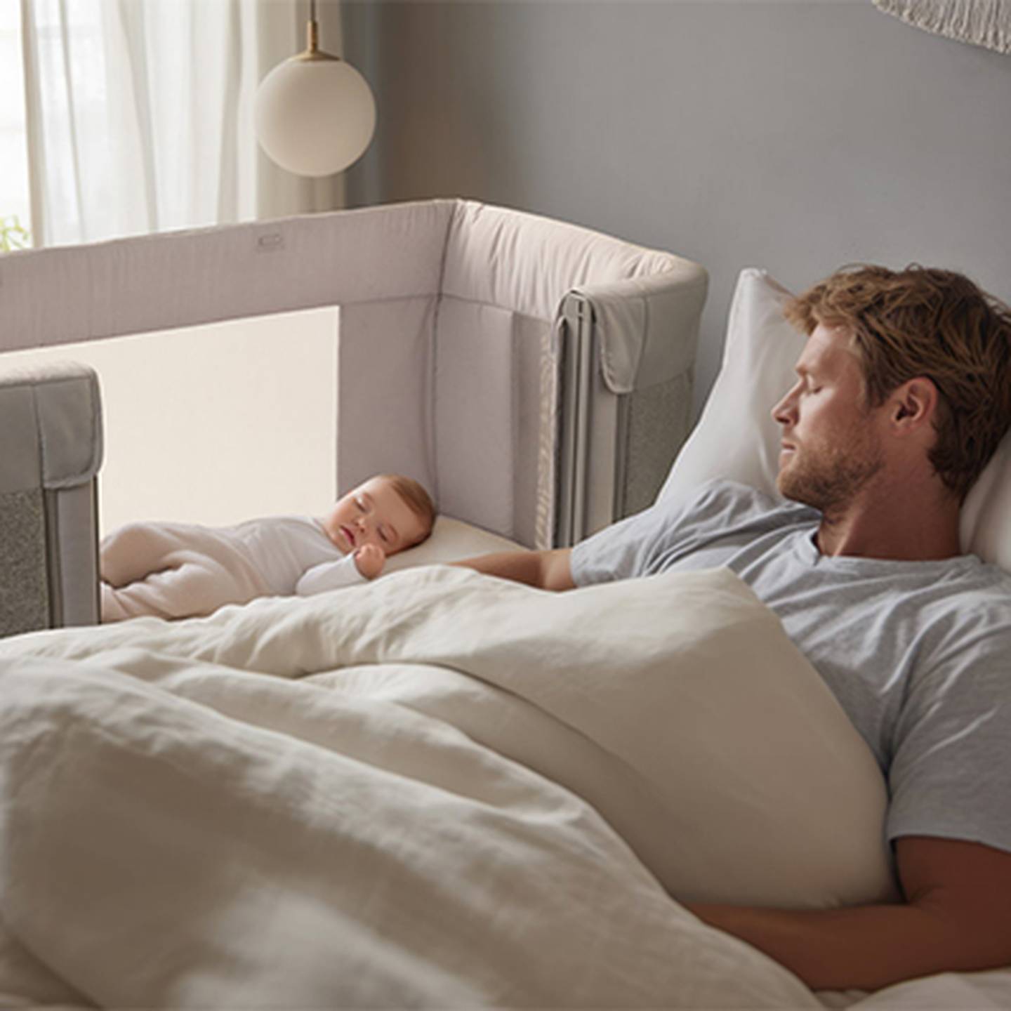 Ein Mann schläft im Bett neben einem Baby in einem Beistellbett. Das Zimmer ist sanft beleuchtet, und beide wirken friedlich und zufrieden.