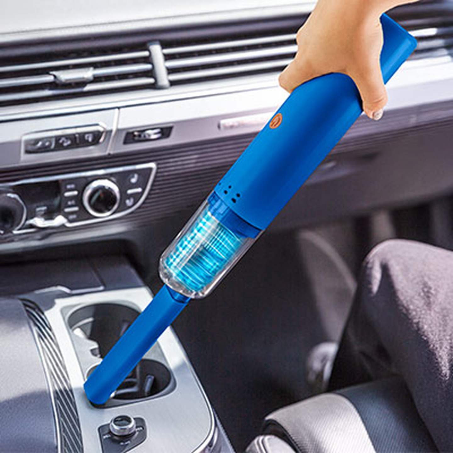 Hand, die mit einem blauen Handstaubsauger den Innenraum eines Autos reinigt, wobei der Fokus auf dem Bereich der Konsole in der Nähe des Schalthebels liegt.