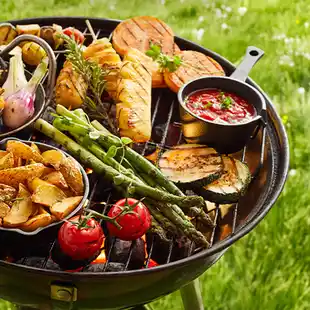 Ein Grill mit Gemüse: Tomaten, Spargel, Kartoffeln, Zucchini, Zwiebeln, Ananas und eine kleine Pfanne mit Soße, draußen auf dem Rasen.