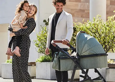 An einem sonnigen Tag steht im Freien eine lächelnde Familie mit einer Frau, die ein Kind hält, neben einem Mann, der einen grünen Kinderwagen schiebt.