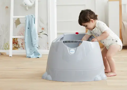Kleinkind im Strampler lehnt sich an eine große graue Plastik-Babytöpfchen-/Trainingswanne auf dem Holzboden des Kinderzimmers.