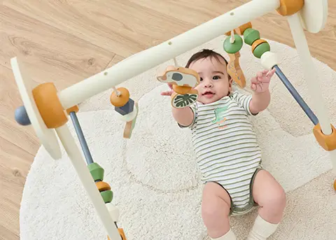Ein Baby liegt auf einem Teppich und greift nach hölzernen Hängespielzeugen an einem Spielbogen. Es trägt einen grün gestreiften Strampler.