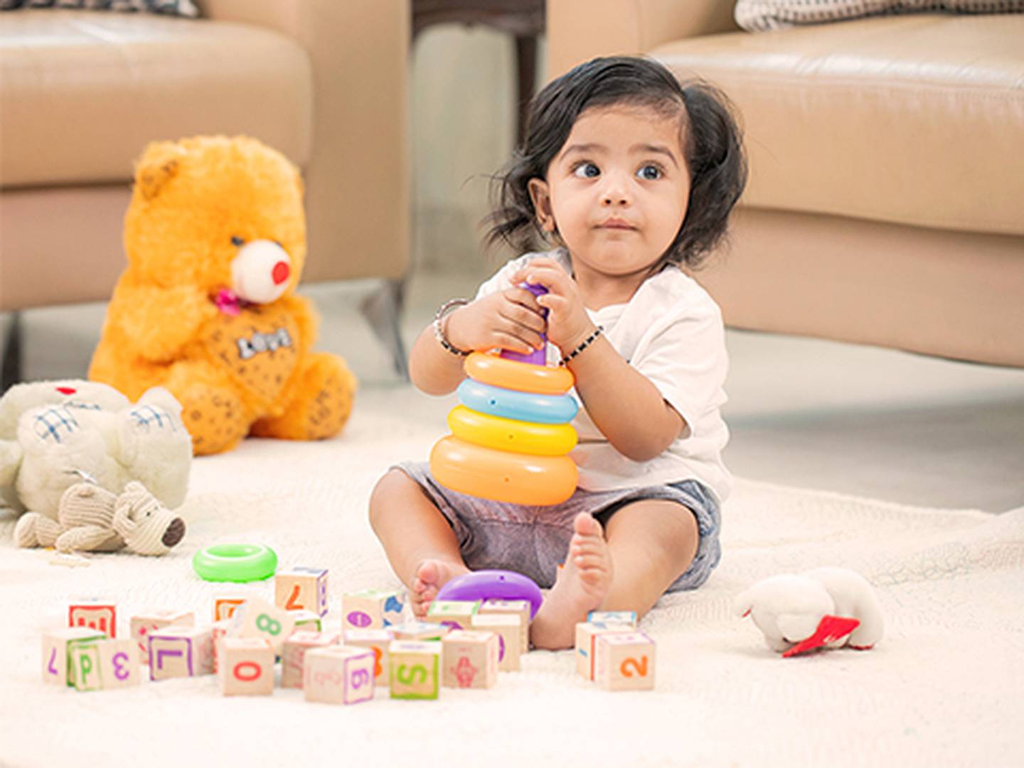 Ein Kleinkind spielt mit bunten Stapelringen, umgeben von Bauklötzen und Plüschtieren auf einem hellen Teppich in einem gemütlichen Wohnzimmer.