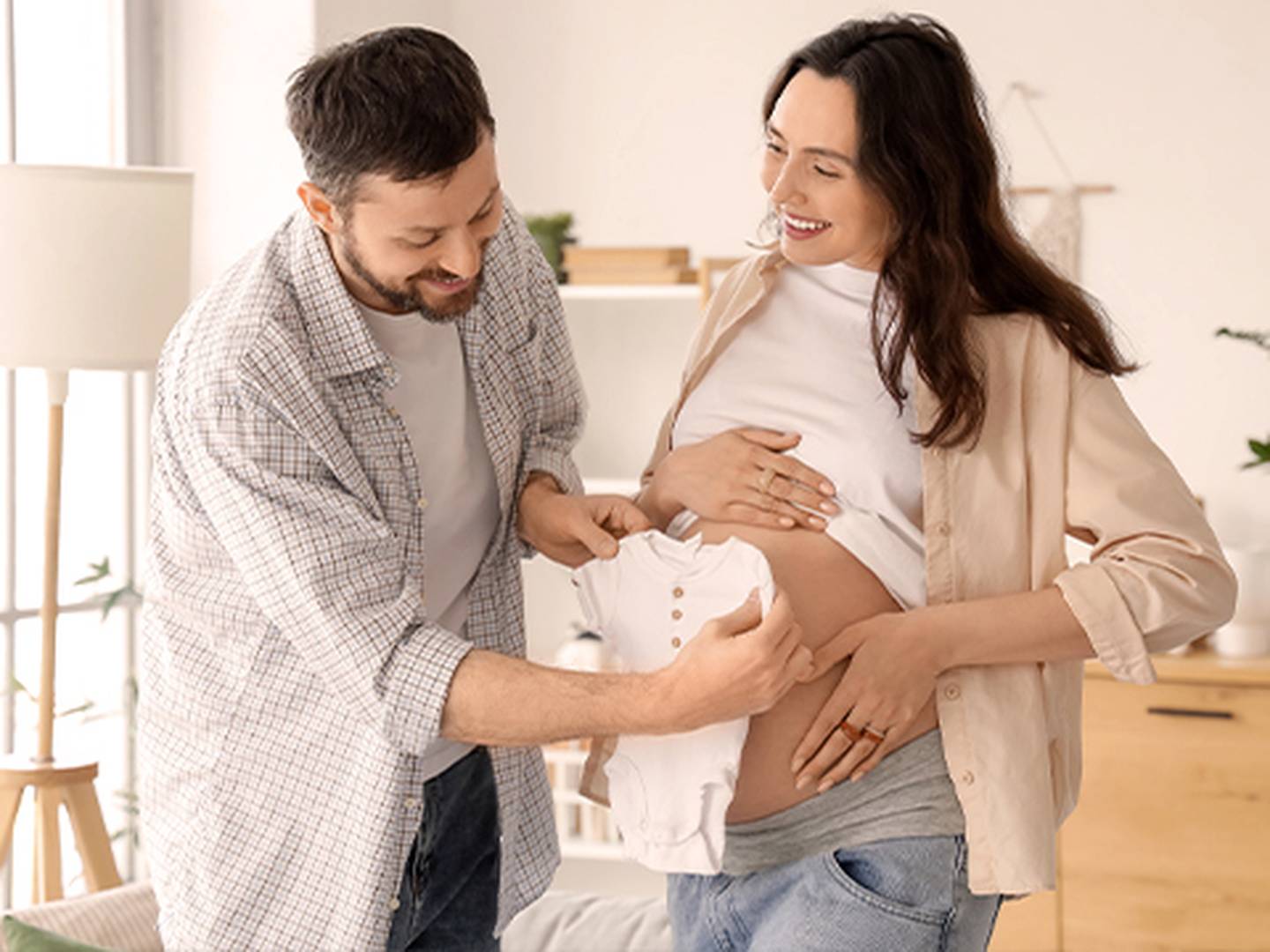 In einem hellen, gemütlichen Wohnzimmer hält der Partner einer lächelnden Schwangeren einen Babybody an ihren Bauch.