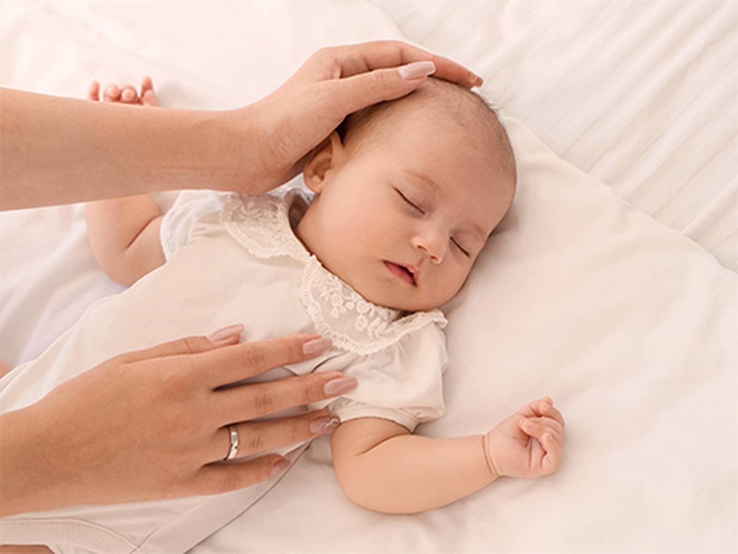 Ein friedlich schlafendes Baby schläft in einem weißen Bettchen, das mit Spitze besetzt ist. Eine Erwachsenenhand stützt sanft den Kopf und ruht auf der Brust.