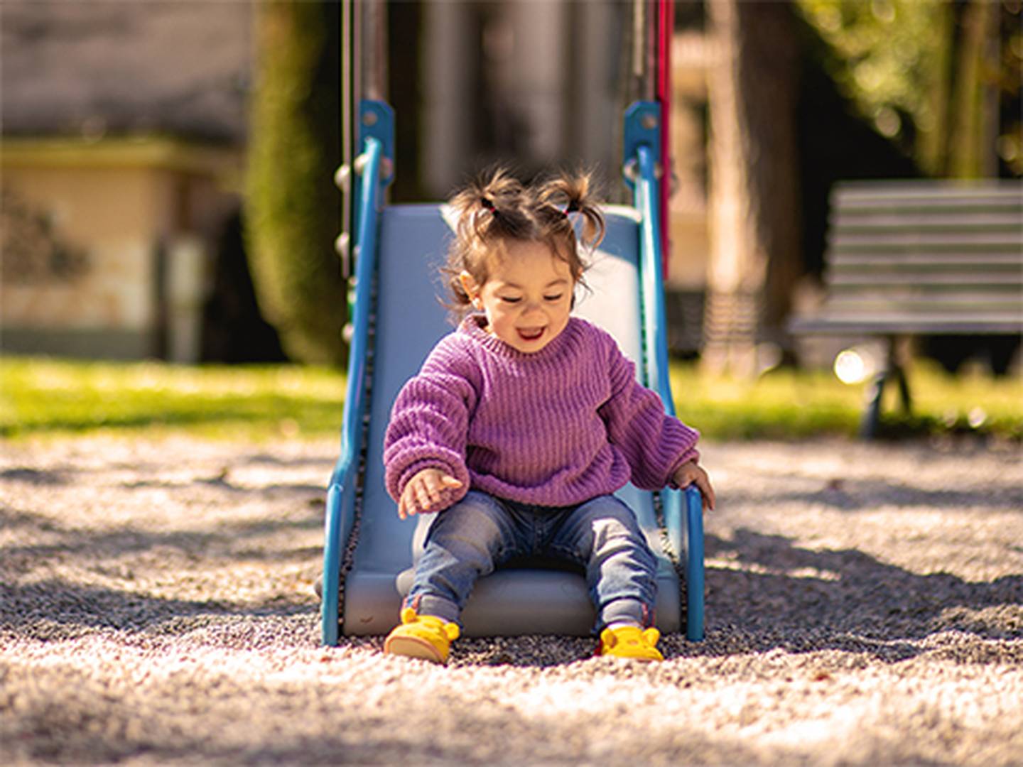 Ein kleines Kind mit Zöpfen und einem lila Pullover rutscht lächelnd und sichtlich vergnügt eine blaue Rutsche auf einem sonnigen Spielplatz hinunter.