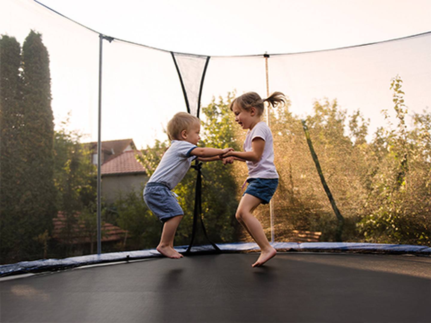 Zwei Kinder spielen vergnügt und halten Händchen auf einem Trampolin in einem Hinterhof, umgeben von Bäumen und Häusern unter dem Abendhimmel.