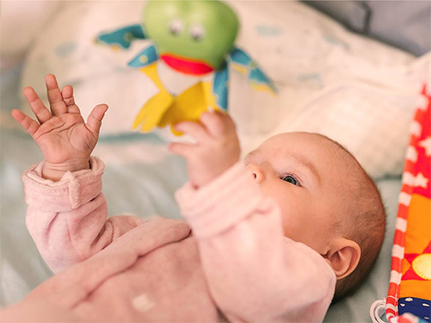 Ein Baby in einem rosa Strampler liegt da und greift nach einem grünen Stofftier darüber; daneben befinden sich ein Kissen und eine gemusterte Decke.