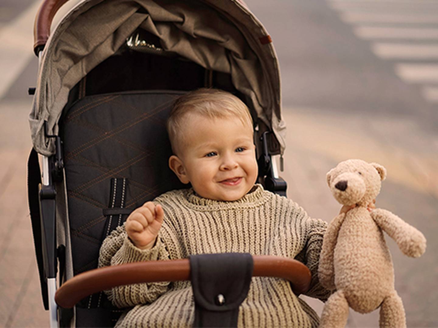 Ein lächelndes Kleinkind in einem Kinderwagen hält einen Teddybären im Arm; es trägt einen Strickpullover. Die Szene spielt sich draußen auf der Straße ab.