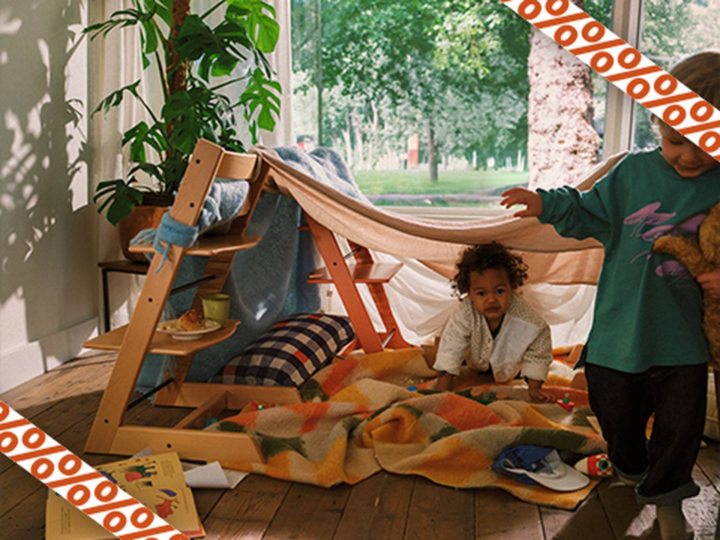 Kinder spielen in einem gemütlichen, aus Decken und einem Holzrahmen gefertigten Zelt im Innenbereich, mit Spielzeug und Kissen auf dem Boden, in der Nähe eines großen Fensters.