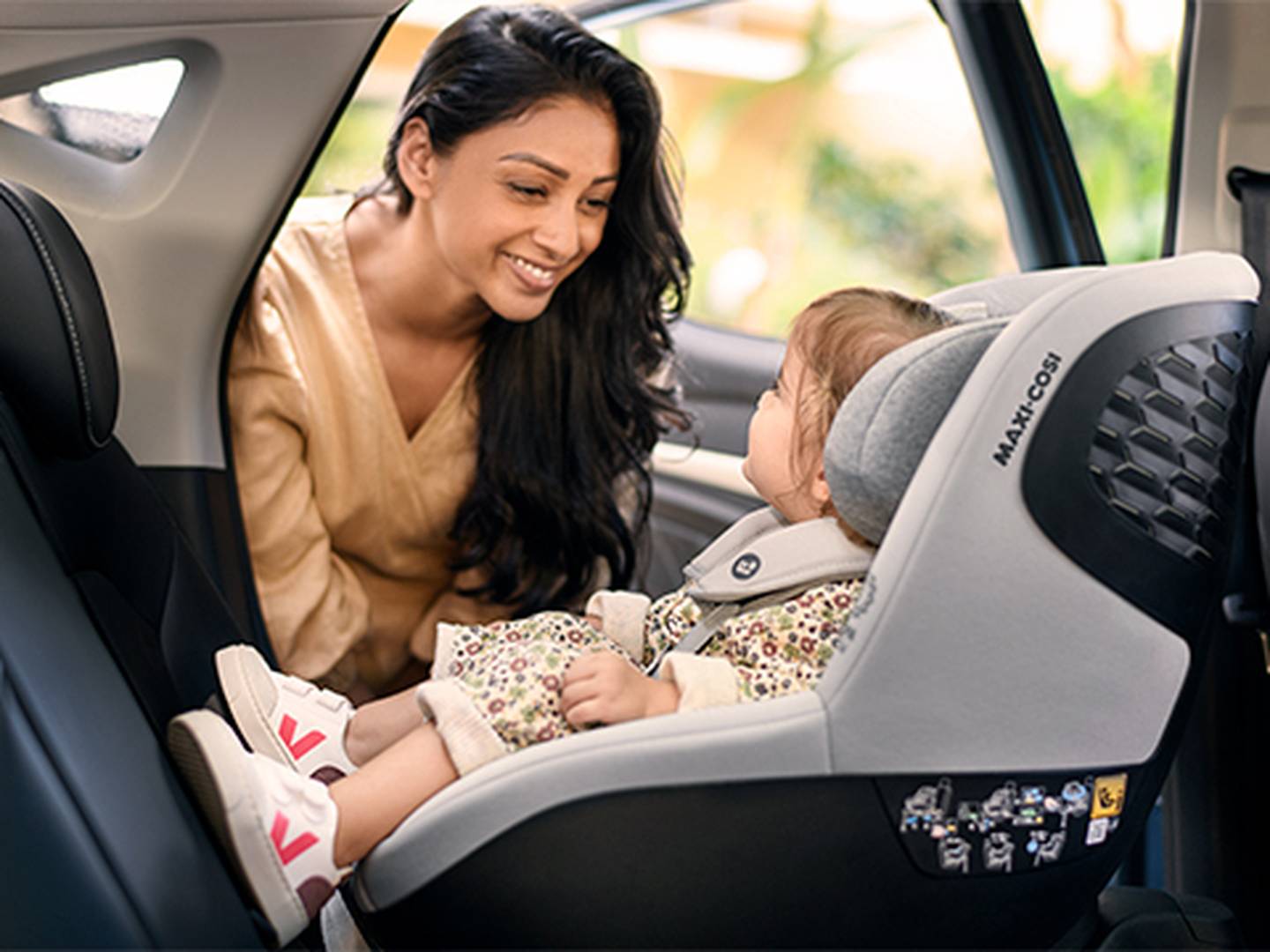 Eine Frau lächelt ein Kleinkind an, das in einem Kindersitz im Auto sitzt. Das Kleinkind trägt Kleidung mit Blumenmuster und weiße Schuhe.