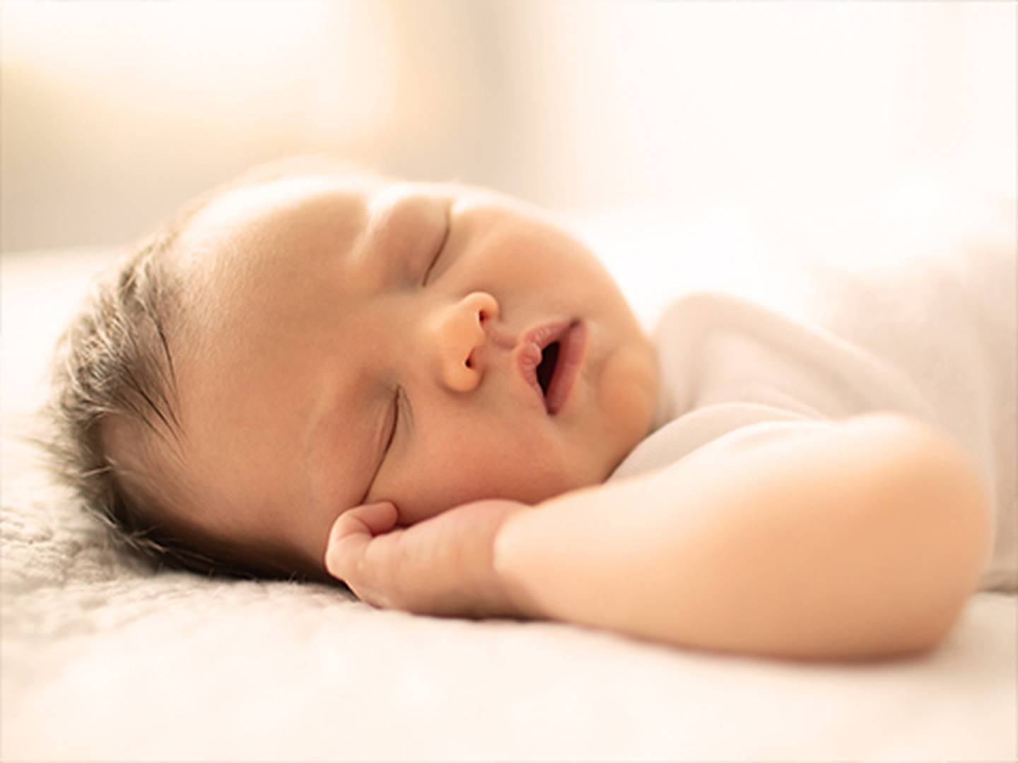 Ein schlafendes Baby liegt mit friedlichem Gesichtsausdruck auf einer weichen Unterlage, bekleidet mit einem weißen Outfit, während sanftes Licht die Szene erhellt.