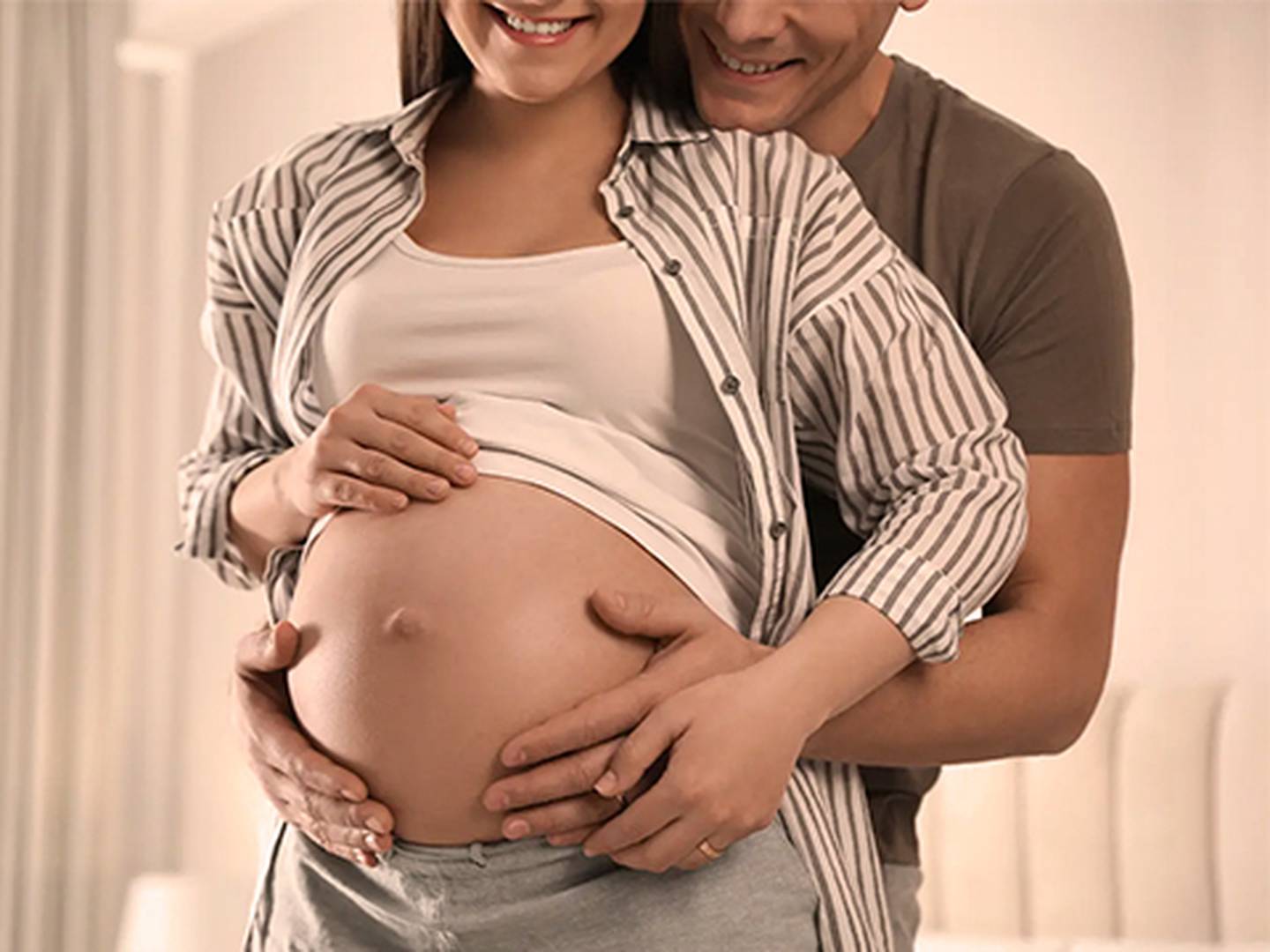 Lächelndes Paar umarmt sich, die Hände sanft auf den schwangeren Bauch der Frau gelegt, in einem sanft beleuchteten Raum.