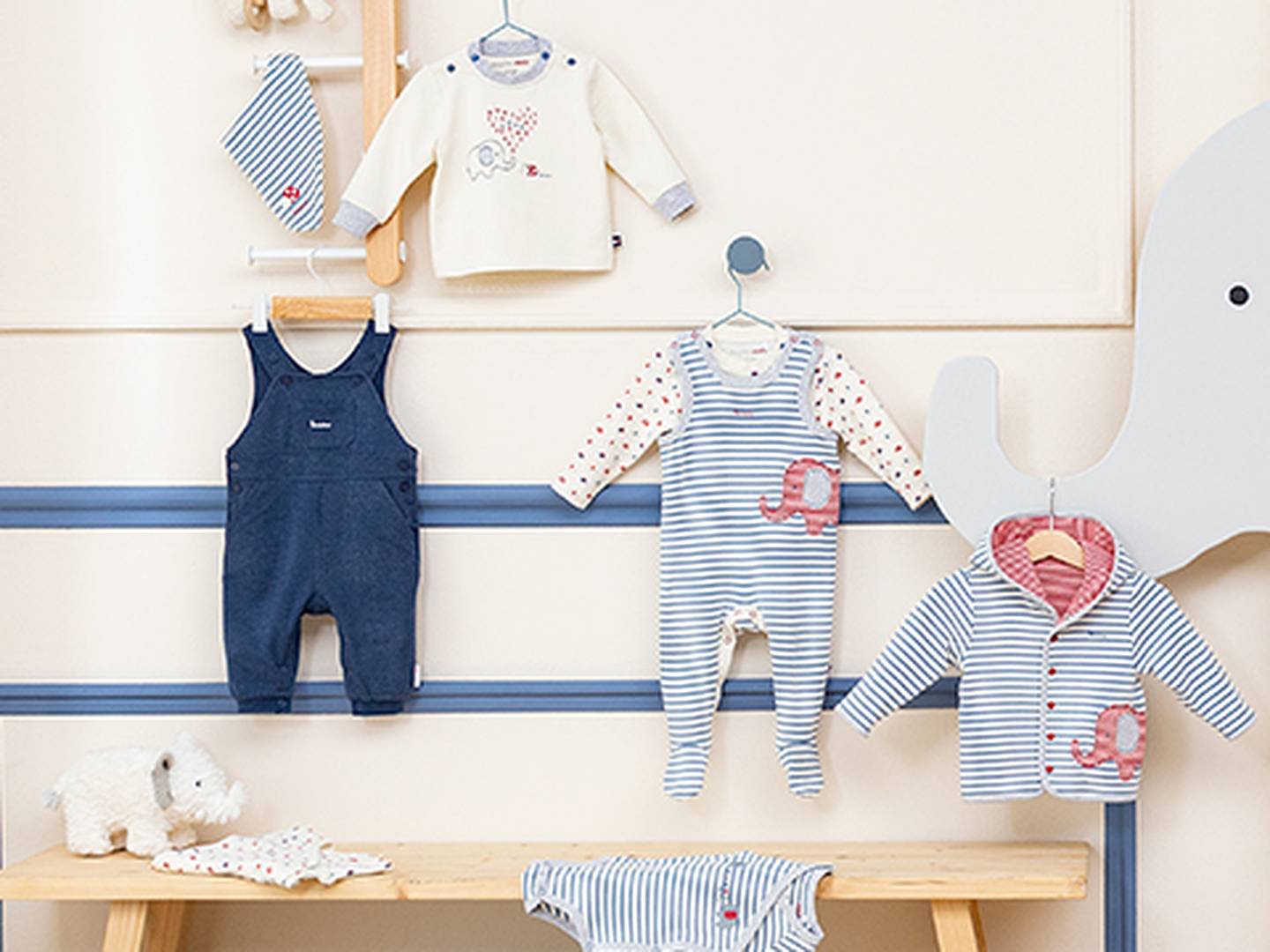 Babykleidungsständer mit gestreiften und einfarbigen Outfits mit Elefantenmotiven. Enthält Strampler, Jacken und Overalls auf einer Holzbank und Wandhaken.