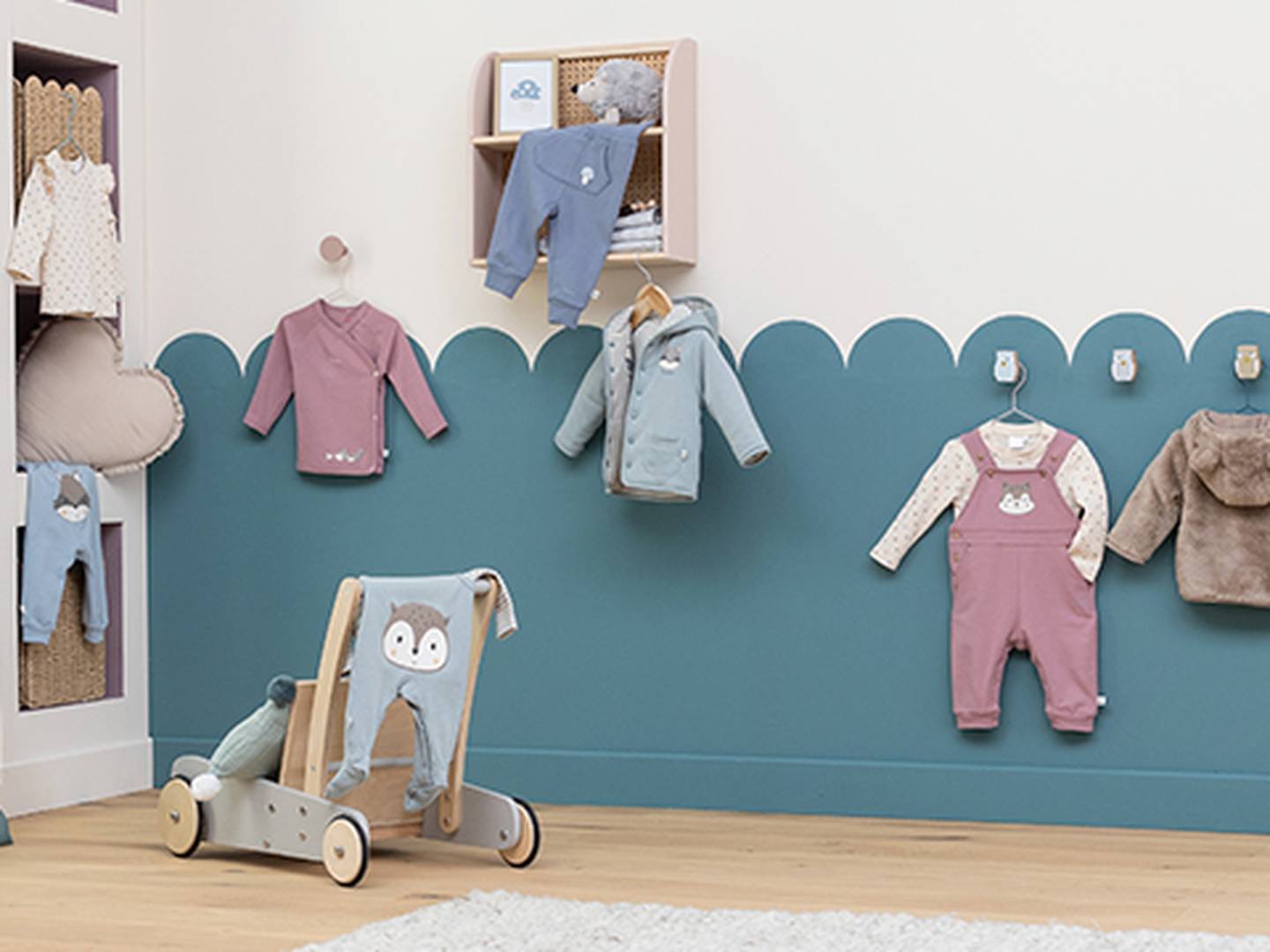 Ein Kinderzimmer mit pastellfarbener Babykleidung, die an einer gewellten blauen Wand hängt, einem Lauflernwagen aus Holz und einem weichen Teppich auf dem Holzboden.