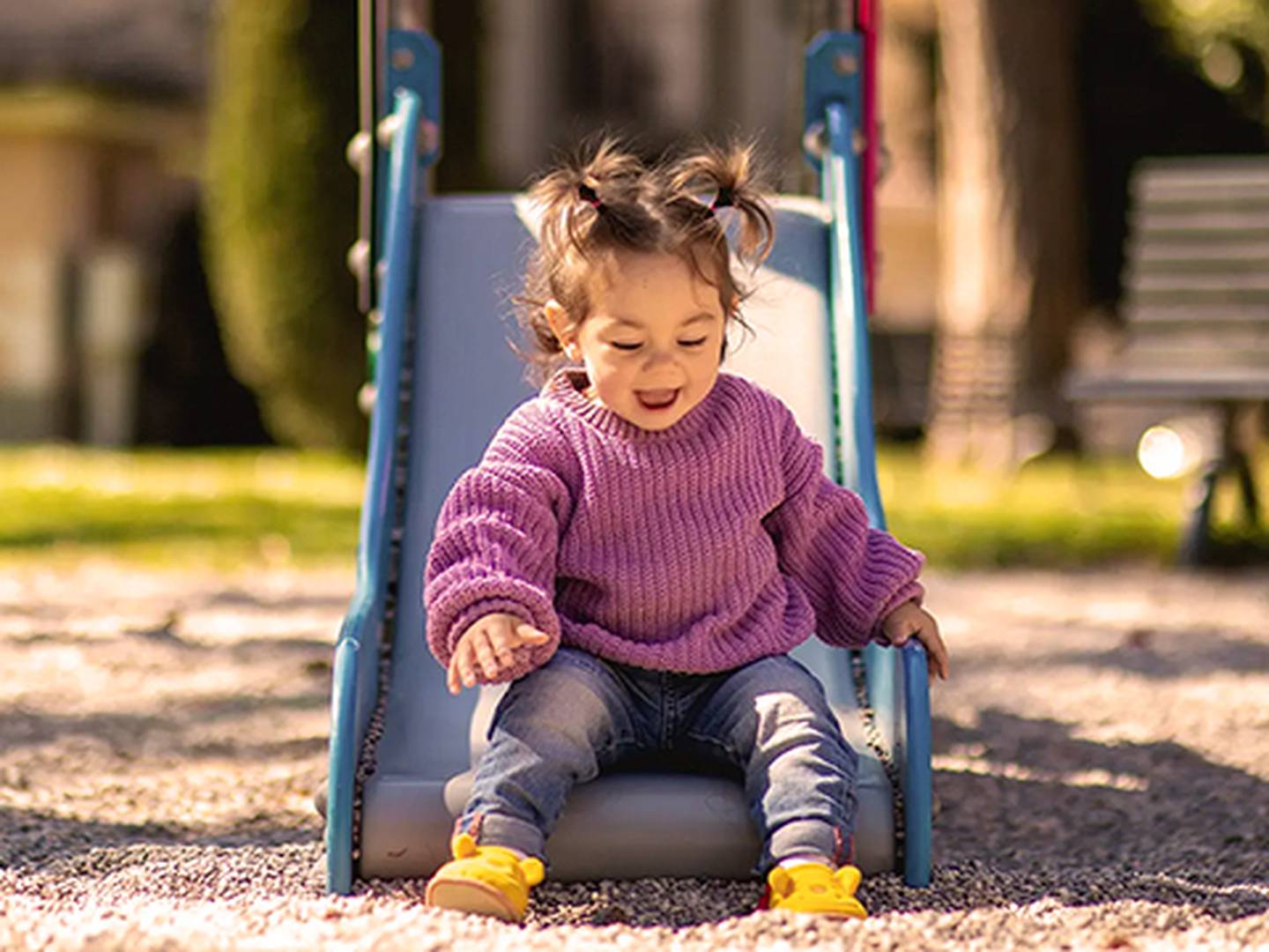 Ein fröhliches Kleinkind mit Zöpfen, das einen lila Pullover und Jeans trägt, rutscht an einem sonnigen Tag eine Rutsche auf einem Spielplatz hinunter.