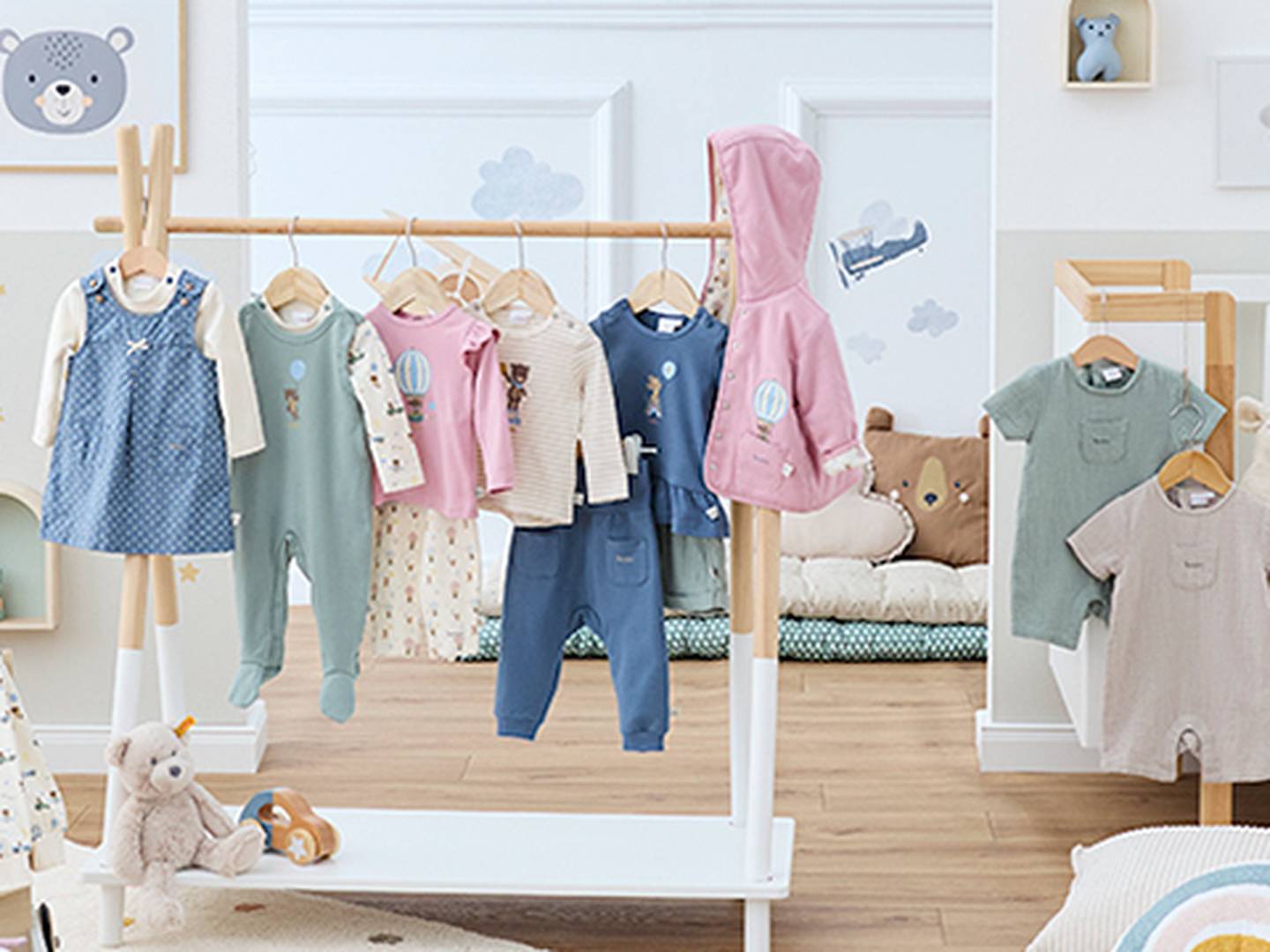 Ein Kinderzimmer mit Kinderkleidung auf einem Holzständer. Die Kleidung umfasst Strampler, Overalls und Jacken in Pastellfarben. Stofftiere liegen auf dem Boden.