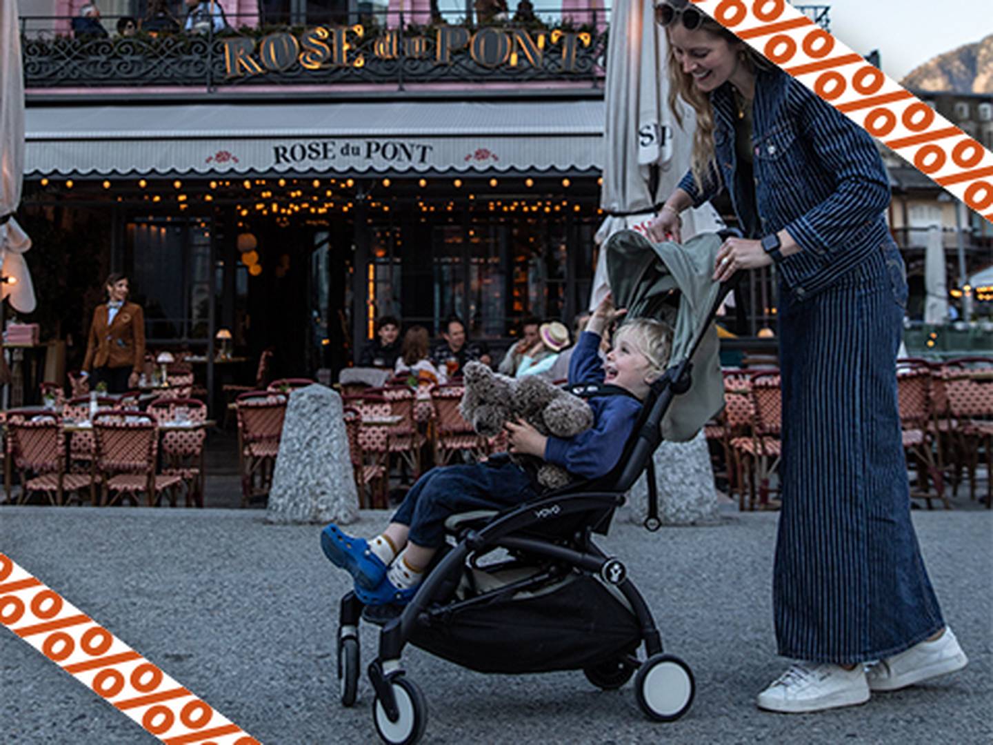 Eine Frau in blauem Outfit beugt sich über einen Kinderwagen und justiert das Verdeck für ein lächelndes Kind, das ein Stofftier hält. Im Hintergrund ist ein Café mit Außenterrasse zu sehen.