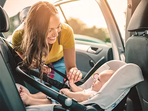 Eine Frau lächelt einem Baby in einem Autositz zu. Sonnenlicht fällt durch die offene Tür und schafft eine warme Atmosphäre.