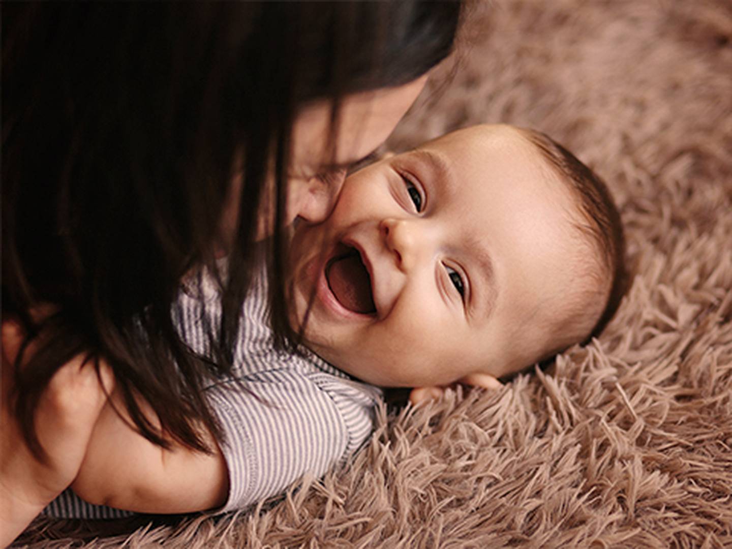 Ein fröhliches Baby liegt auf einem weichen, strukturierten Teppich, lächelt und wird von einer Person mit dunklem Haar geknuddelt.