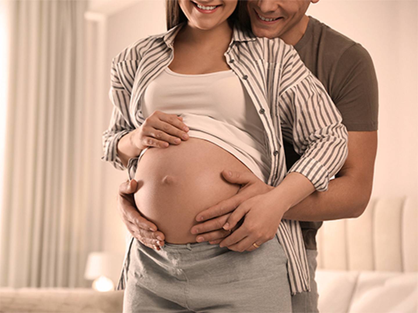 Ein Paar umarmt sich liebevoll, die Hände des Mannes ruhen auf dem schwangeren Bauch der Frau – eine Atmosphäre der Wärme und Vorfreude in einem gemütlichen Zimmer.
