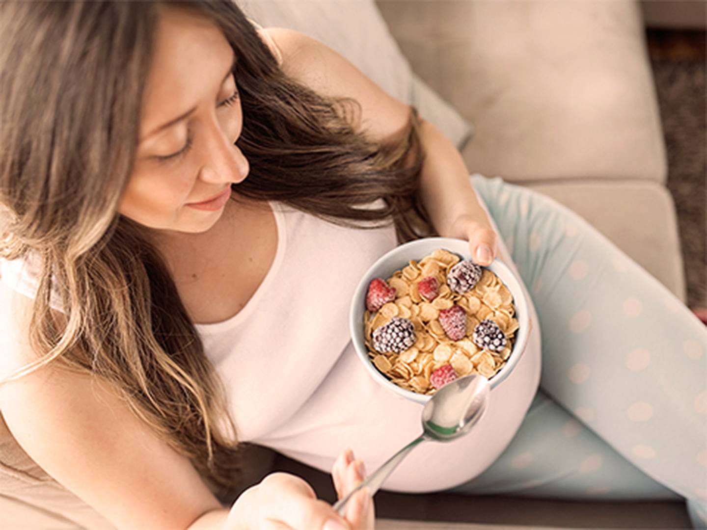 Schwangere Frau auf einer Couch, die eine Schüssel Müsli mit Brombeeren und Himbeeren hält, bekleidet mit einem weißen Tanktop und hellblauen Hosen.