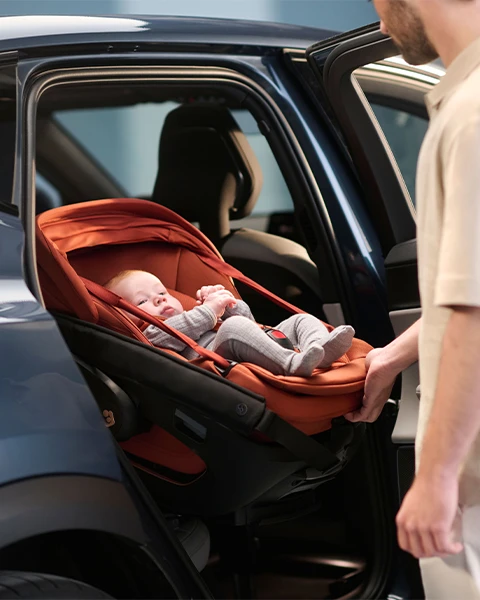Eine Person setzt ein Baby in einem roten Kindersitz in ein Auto. Das Baby trägt graue Kleidung und wirkt entspannt und geborgen.