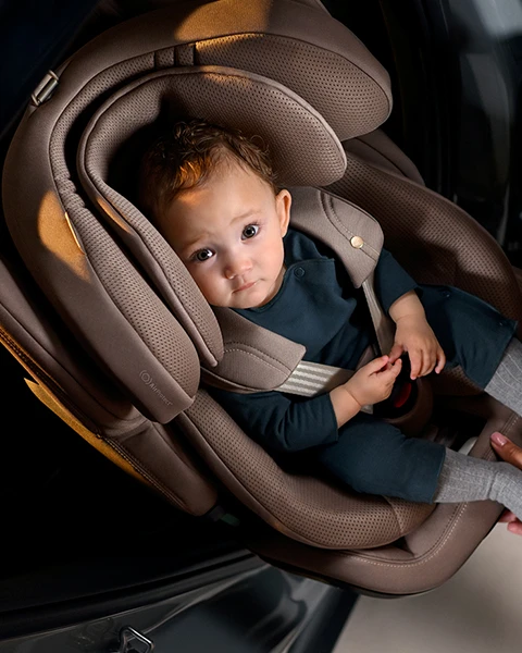 Ein Baby in einem Kindersitz blickt zu einem Erwachsenen auf, der seinen Fuß hält. Der Kindersitz ist auf dem Rücksitz eines Fahrzeugs installiert.