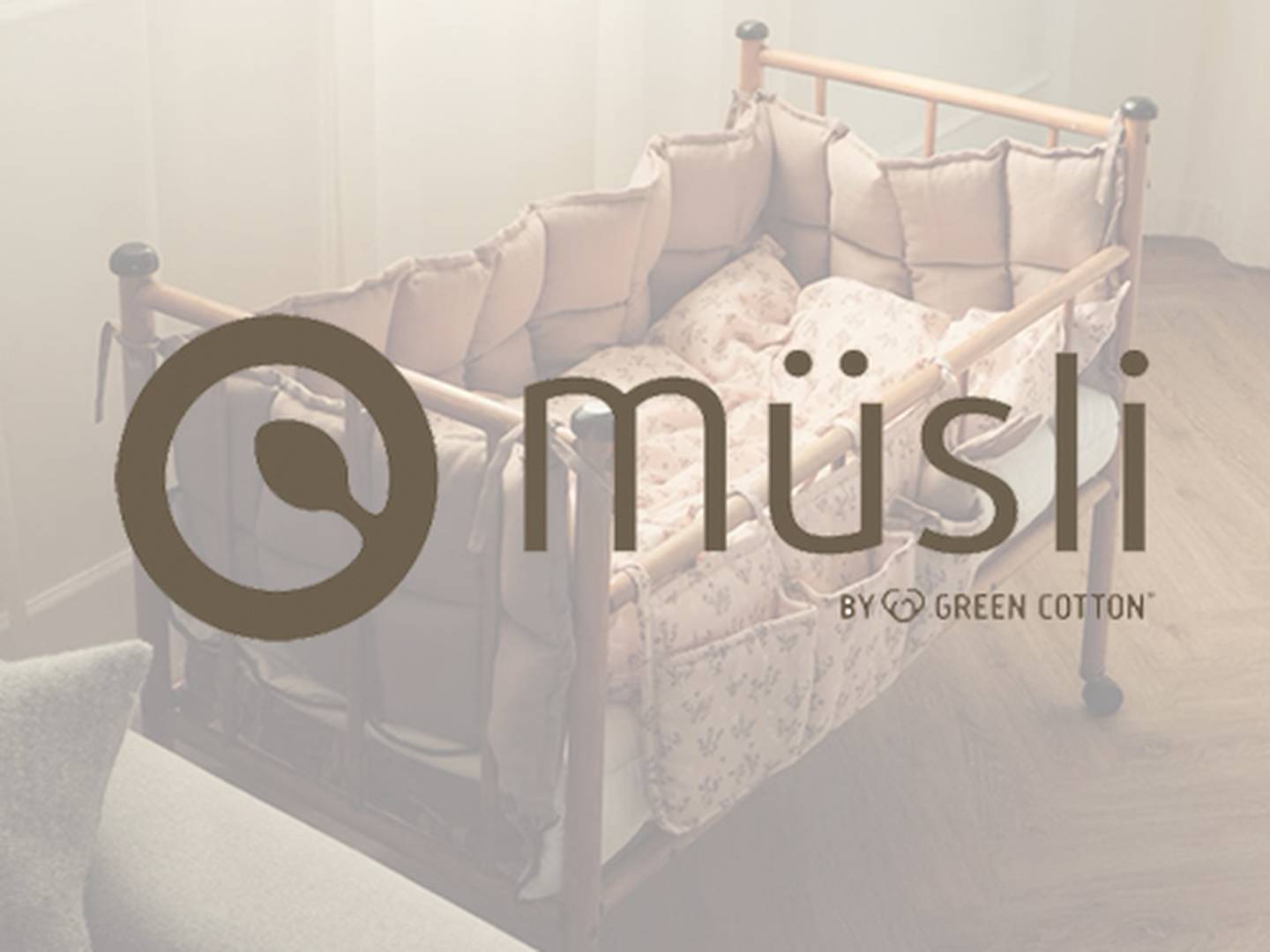 Ein hölzernes Babybett mit beiger Polsterung und rosa Decke in einem sanft beleuchteten Zimmer. Darüber prangt das Logo „Müsli by Green Cotton“.