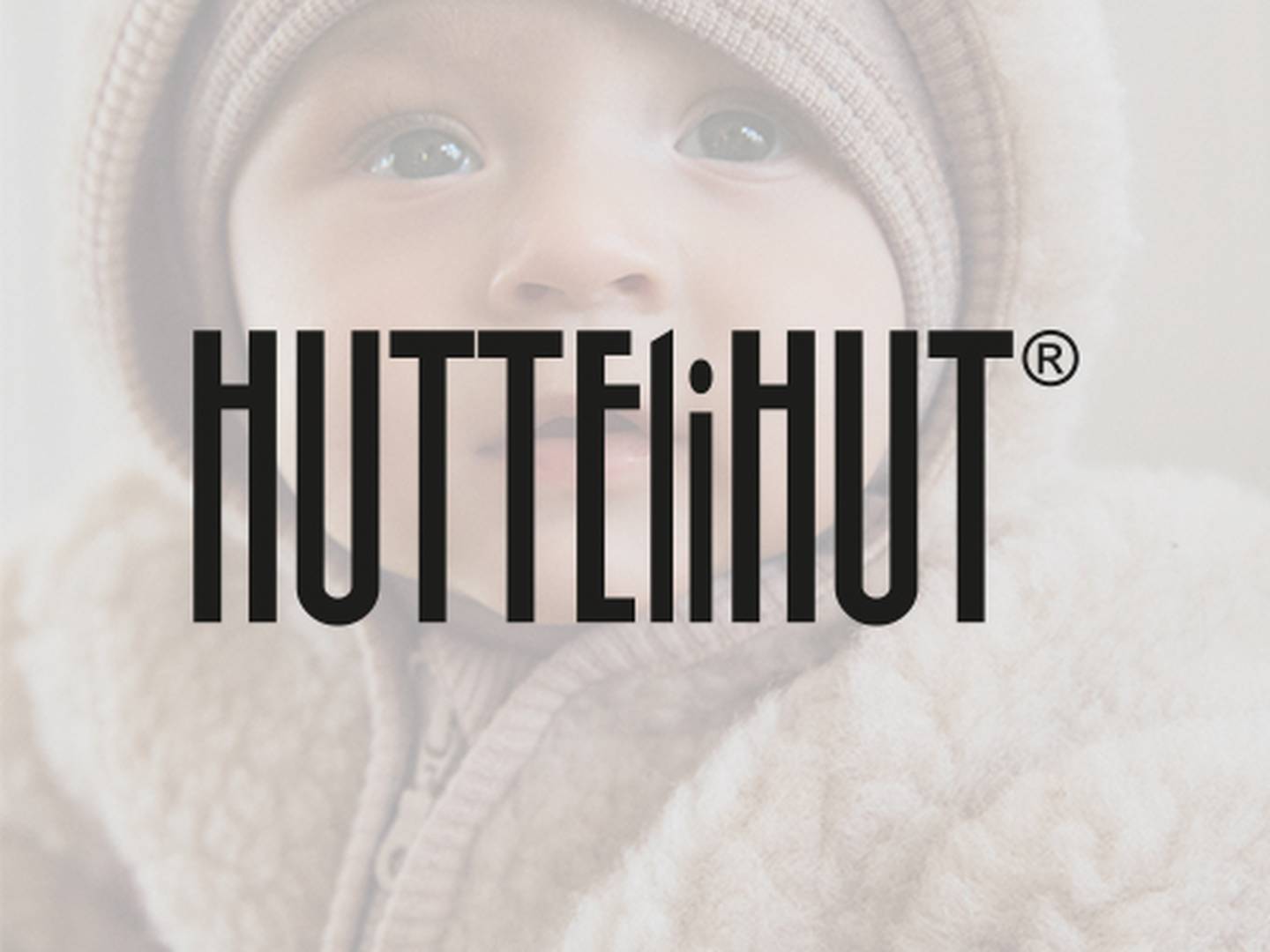 Ein Baby in einem gemütlichen, beigen Outfit und passender Mütze, über das der Text „HUTTEliHUT“ gelegt ist.