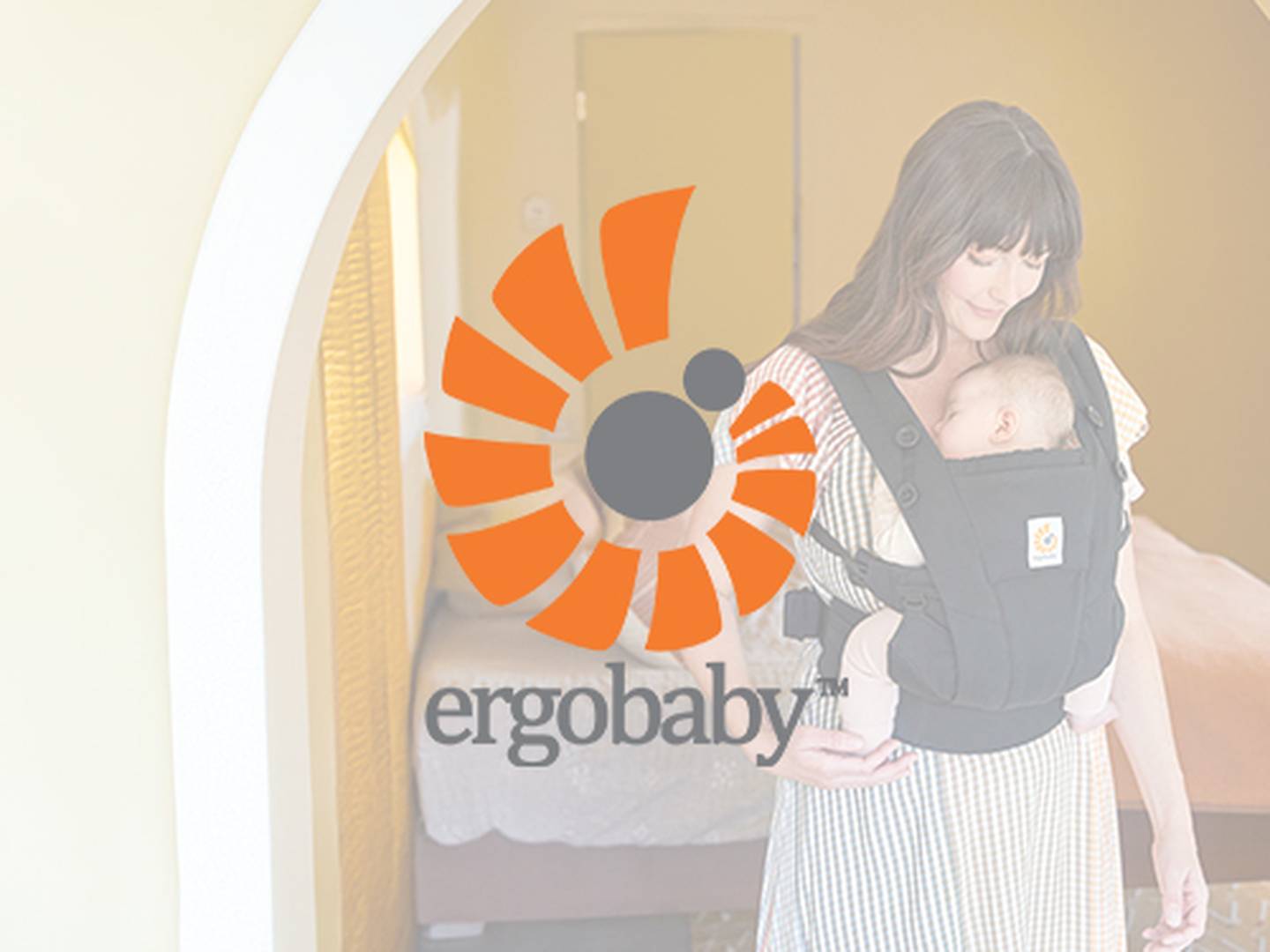 Frau hält ein schlafendes Baby in einer schwarzen Babytrage; sie steht in einem sanft beleuchteten Raum mit dem Ergobaby-Logo.