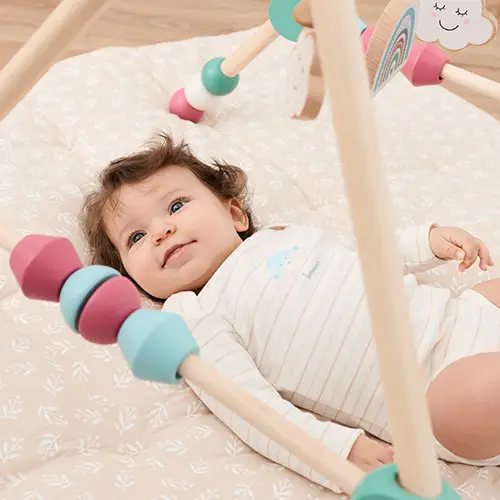 Ein Baby liegt auf einer weichen Decke unter einem hölzernen Spielbogen mit bunten Perlen und wirkt zufrieden und vertieft in sein Spiel.