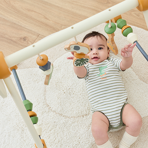 Ein Baby liegt auf einem Teppich und greift nach hölzernen Hängespielzeugen an einem Spielbogen. Es trägt einen grün gestreiften Strampler.