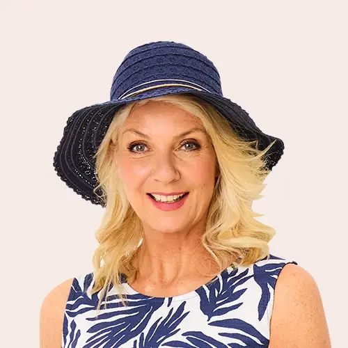 Lächelnde Frau mit langen blonden Haaren, die einen marineblauen Sonnenhut und ein weißes Kleid mit blauen Blattmustern trägt, posiert vor einem einfarbigen Hintergrund.