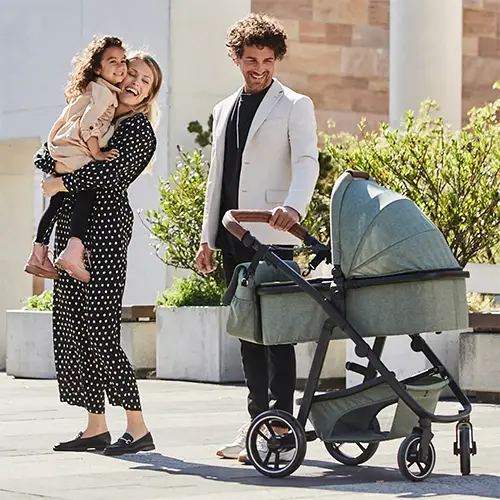 Eine lächelnde Frau hält ein Kind, während sie an einem sonnigen Tag in einer städtischen Umgebung neben einem Mann mit einem Kinderwagen steht.