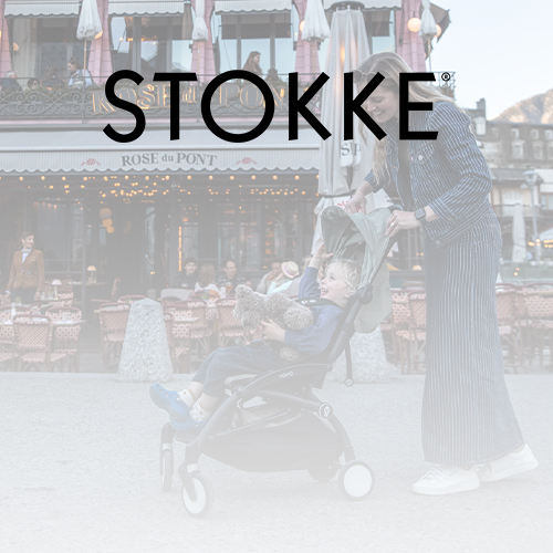 Eine Person schiebt einen Kinderwagen mit einem Kind, das einen Teddybären hält, vor einem Café. Großer Schriftzug „STOKKE“ darüber.