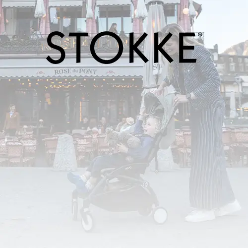 Eine Person schiebt einen Kinderwagen mit einem Kind, das einen Teddybären hält, vor einem Café. Großer Schriftzug „STOKKE“ darüber.