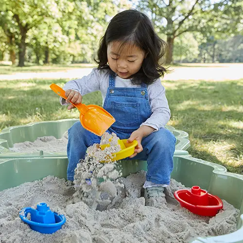 Ein junges Mädchen spielt im Sandkasten und füllt mit einer gelben Schaufel einen grünen Eimer mit Sand. Bunte Spielzeugboote liegen in der Nähe.