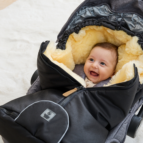 Ein lächelndes Baby, eingehüllt in einen flauschigen, fellgefütterten schwarzen Kinderwagen-Fußsack