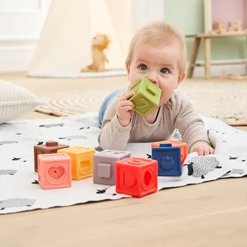 Baby liegt auf einem gemusterten Teppich, spielt mit bunten Spielzeugblöcken und kaut auf einem grünen Block. Ein gemütliches Zimmer mit weicher Beleuchtung im Hintergrund.