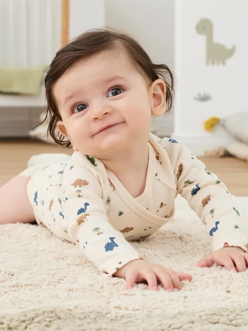 Ein Baby in einem Dinosaurier-Strampler krabbelt lächelnd auf einem weichen Teppich. Im Hintergrund ist ein Dinosaurier-Wandtattoo zu sehen.