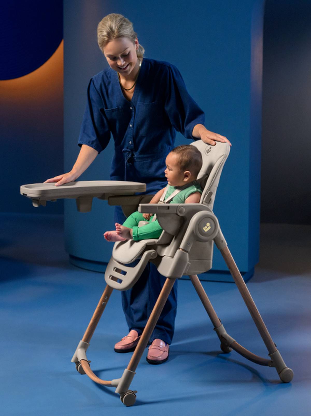 Eine Frau in einem blauen Outfit justiert das Tablett eines Hochstuhls, auf dem ein Baby in Grün sitzt, vor einem modernen, sanft beleuchteten Hintergrund.