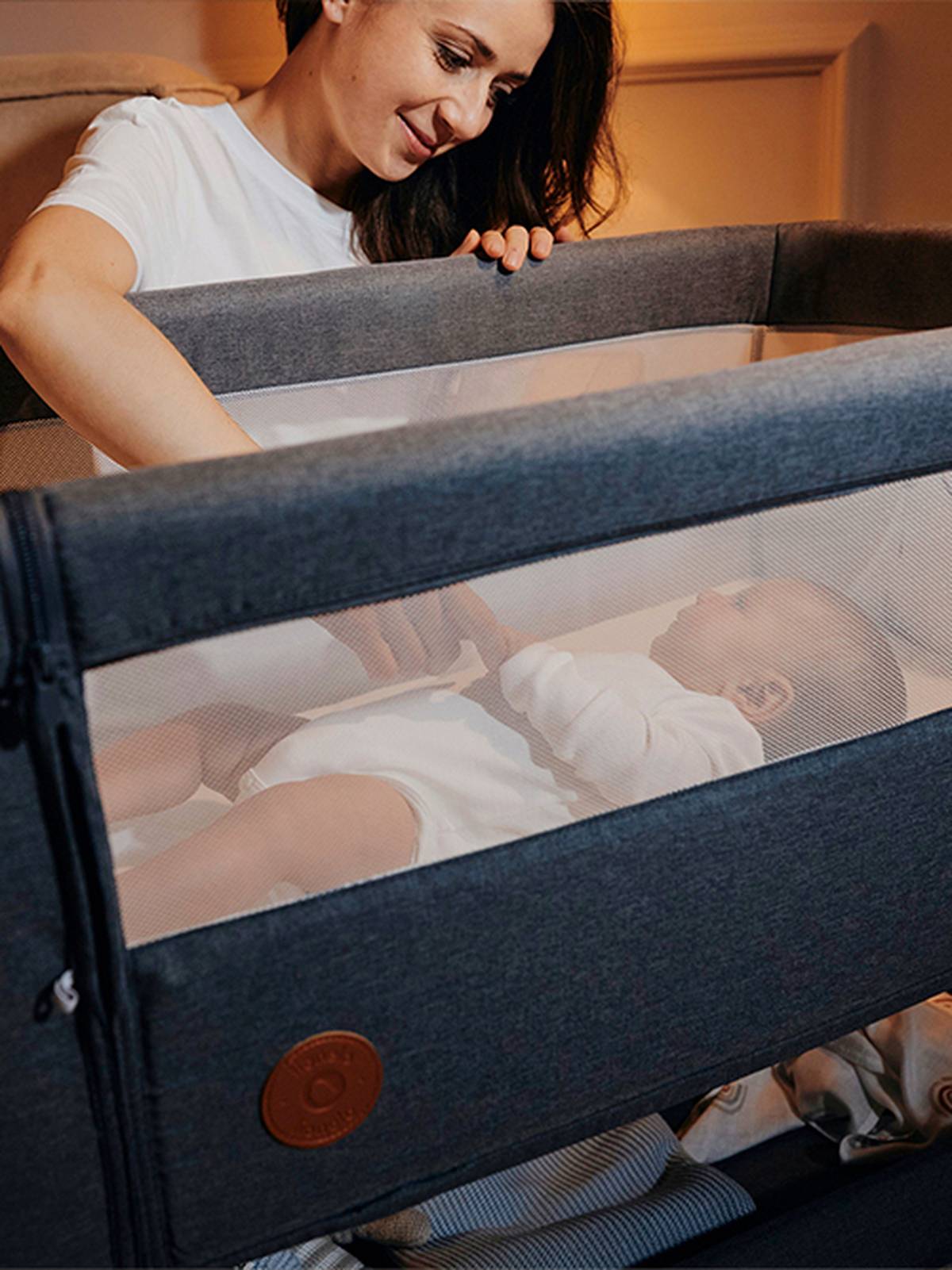 Eine Frau kümmert sich sanft um ein Baby, das in einem grauen Kinderbett mit Netzseiten liegt; beide sind in Weiß gekleidet. Der Raum ist warm beleuchtet.