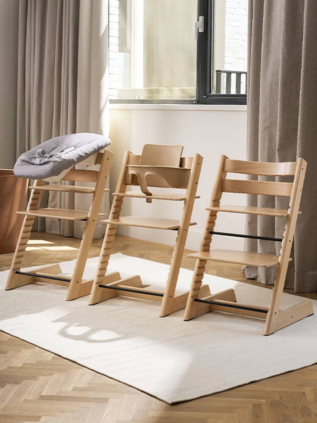 Drei hölzerne Hochstühle, jeder mit unterschiedlichem Sitzzubehör, stehen auf einem hellen Teppich in einem sonnendurchfluteten Zimmer mit Vorhängen und Fenster.