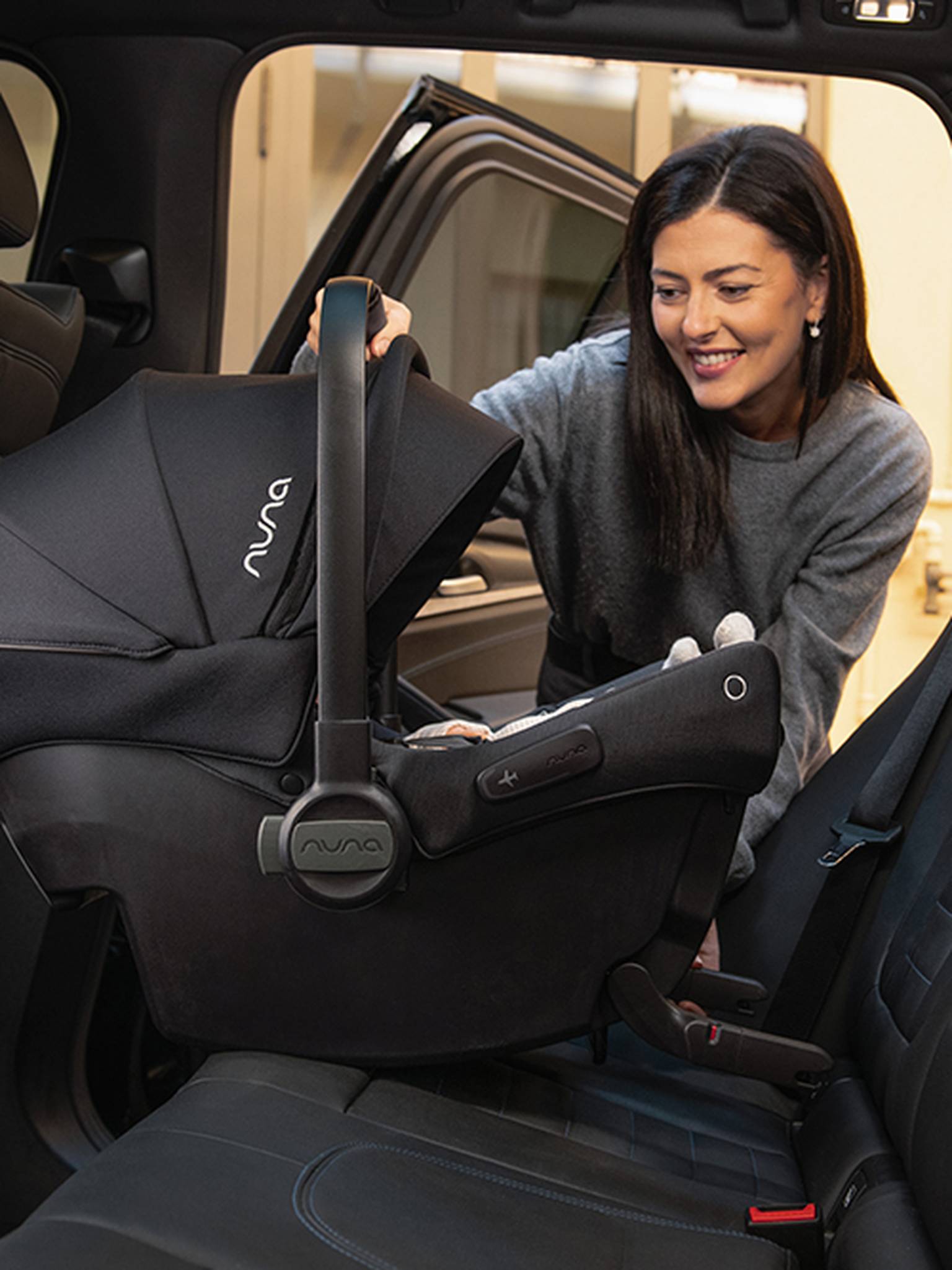 Eine Frau installiert lächelnd einen schwarzen Nuna-Babysitz auf dem Rücksitz eines Fahrzeugs.