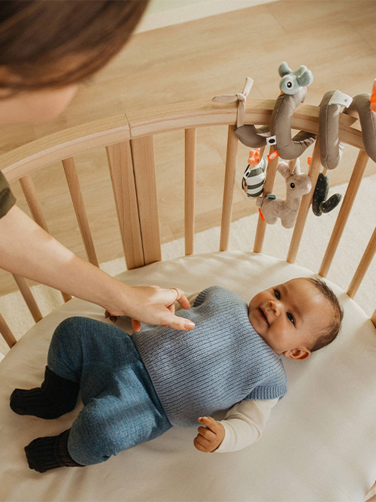 Ein lächelndes Baby liegt in einem Kinderbett, gekleidet in einen blauen Pullover und eine blaue Hose, eine Hand streckt sich nach ihm aus und darüber hängen Plüschtiere.