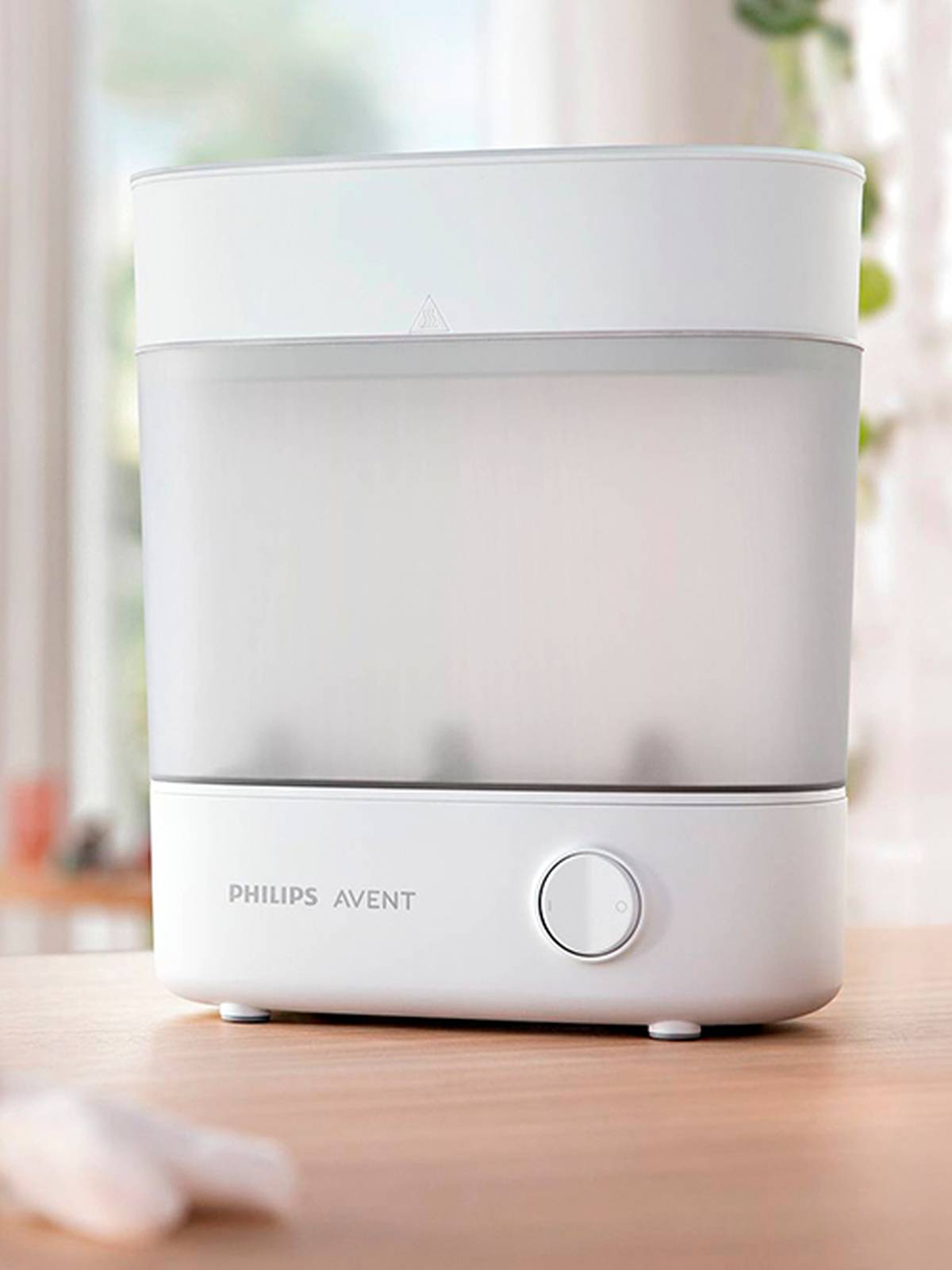 Weißer Philips Avent Sterilisator auf einer Holzoberfläche, mit elegantem Design und rundem Knopf, aufgestellt in einem Innenraum mit sanftem, durch ein Fenster einfallendem Licht.