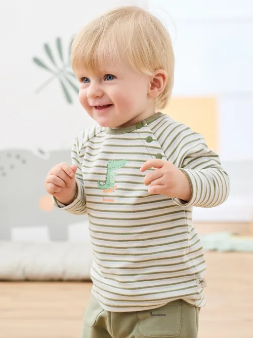 Lächelndes Kleinkind in einem gestreiften Shirt mit Dinosaurier-Aufdruck, das drinnen auf einem Holzboden steht. Der Hintergrund zeigt eine sanfte, neutrale Einrichtung.