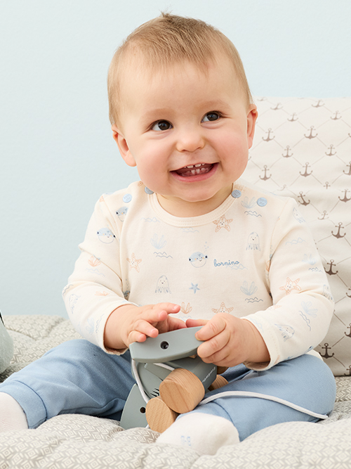 Ein lächelndes Baby in einem gemusterten Strampler und blauen Hosen sitzt auf einem Bett und hält ein Holzspielzeug in der Hand.