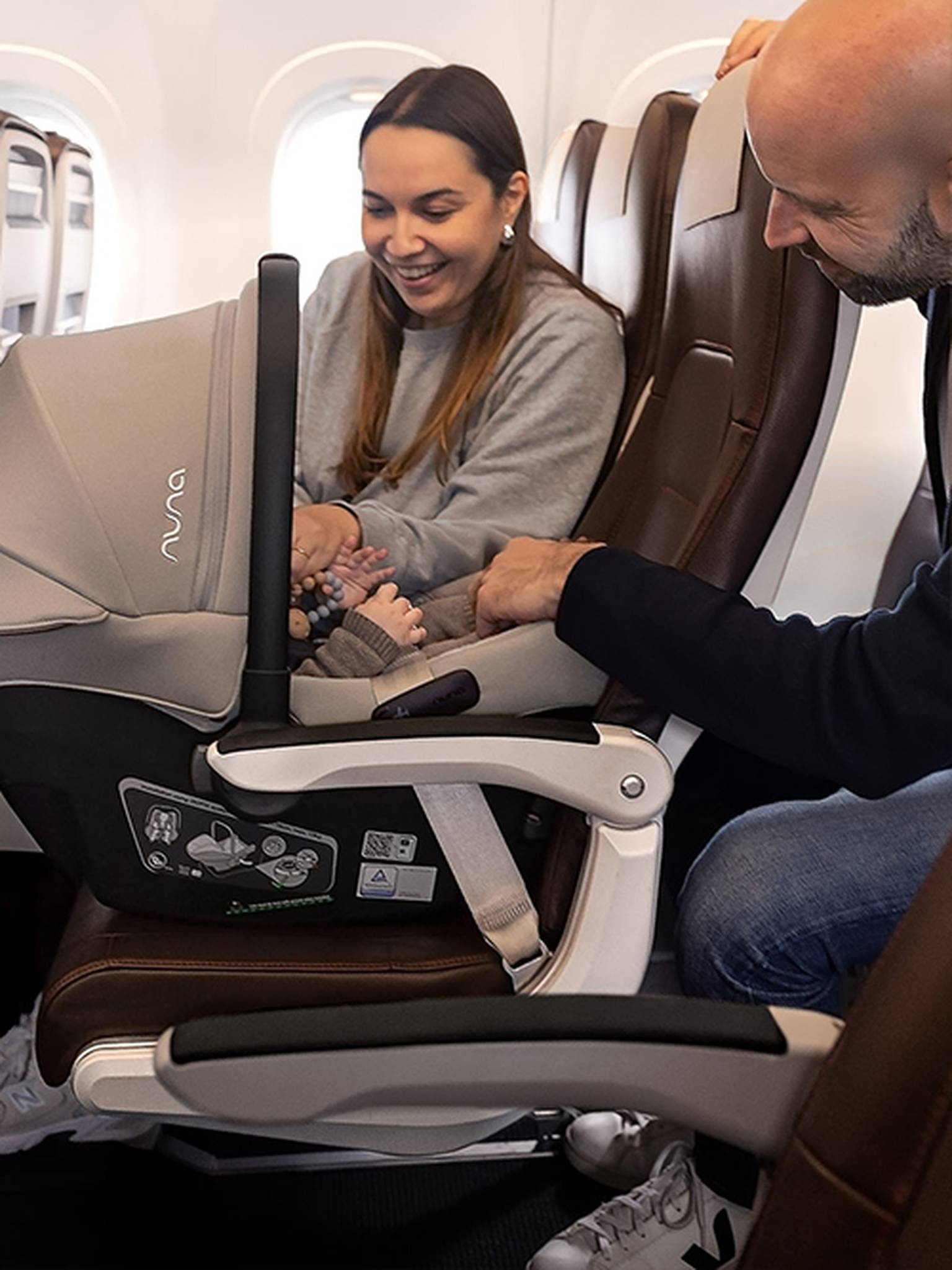 Ein lächelndes Paar interagiert mit einem Baby in einem Kindersitz in einem Flugzeug; im Hintergrund sind braune Ledersitze und ein Fenster zu sehen.
